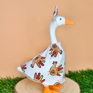 May include: A white goose figurine wearing a white fabric outfit featuring a repeating pattern of brown turkeys with colorful red and yellow tail feathers. The goose has an orange beak and orange feet, and it is wearing a small matching headpiece.