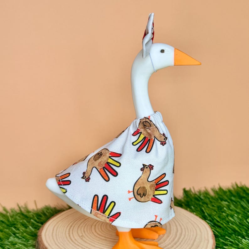 May include: A white goose figurine wearing a white fabric outfit featuring a repeating pattern of brown turkeys with colorful red and yellow tail feathers. The goose has an orange beak and orange feet, and it is wearing a small matching headpiece.