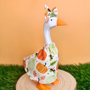 May include: A white plastic goose figurine wearing a fabric dress and a matching bow on its head. The fabric features a pattern of orange pumpkins, green pumpkin outlines, and brown acorns on a white background. The goose has an orange beak and feet.