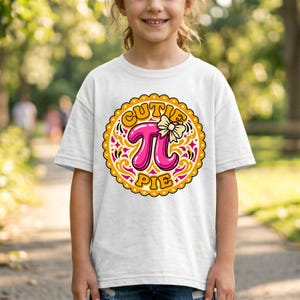 Puede incluir: Camiseta blanca con un diseño gráfico colorido. El diseño incluye las palabras "CUTIE PIE" en una fuente lúdica, un gran símbolo pi rosa y un lazo. El gráfico está dentro de un borde decorativo y festoneado.