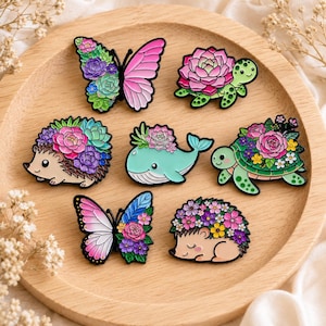 Pode incluir: Um conjunto de sete pins de esmalte com designs de animais e insetos decorados com flores e suculentas coloridas. O conjunto inclui duas borboletas, dois ouriços, duas tartarugas e uma baleia, em tons vibrantes de rosa, verde, azul e roxo.