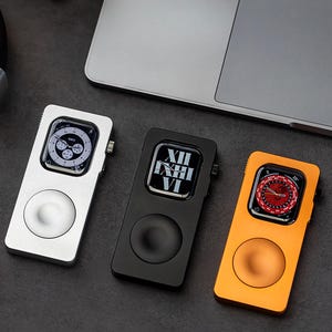 May include: Three metal Apple Watch cases in silver, black, and orange. Each case has a circular button and a screen displaying the watch face. A laptop is visible in the background.