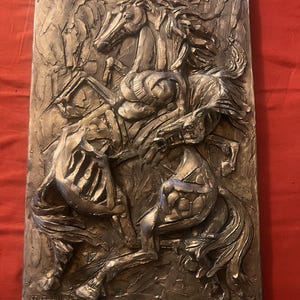 May include: A rectangular, metallic relief sculpture depicting a rearing horse and rider in a dynamic pose. The artwork is rendered in shades of silver and gray, with textured details and visible brushstrokes. The artist's signature is at the bottom.