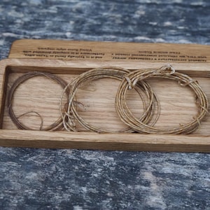 Könnte beinhalten: Ein rechteckiges Holztablett mit heller Oberfläche, das drei aufgewickelte Stränge aus dünner, naturfarbener Kordel enthält. Der obere Rand des Tabletts ist mit eingraviertem Text in Englisch und Kyrillisch versehen.