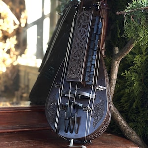 Peut inclure: Une nyckelharpa brun foncé, une vielle à clavier suédoise, avec des motifs sculptés complexes. L'instrument comporte plusieurs cordes, un archet et une manivelle. Il est présenté sur une surface en bois avec un reflet miroir et de la verdure en arrière-plan.