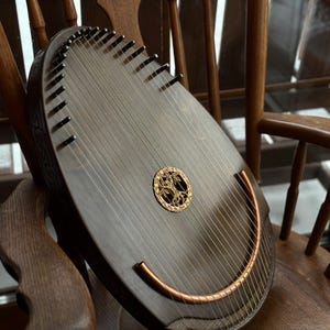May include: A dark wooden stringed instrument with a curved copper bar and a decorative gold-colored emblem. The instrument is resting on a wooden chair, showcasing its intricate design and craftsmanship. The strings are visible, stretched across the body.