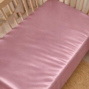 May include: A dusty rose satin crib sheet. The sheet is neatly placed on a wooden crib frame. The crib is empty, and the sheet is the focal point. A woven mat is visible in the lower right corner.