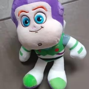 May include: A plush toy of the character Buzz Lightyear from the Toy Story franchise. The toy has a purple helmet, a white face, and a green and white spacesuit. The eyes are large and blue, and the toy is smiling.