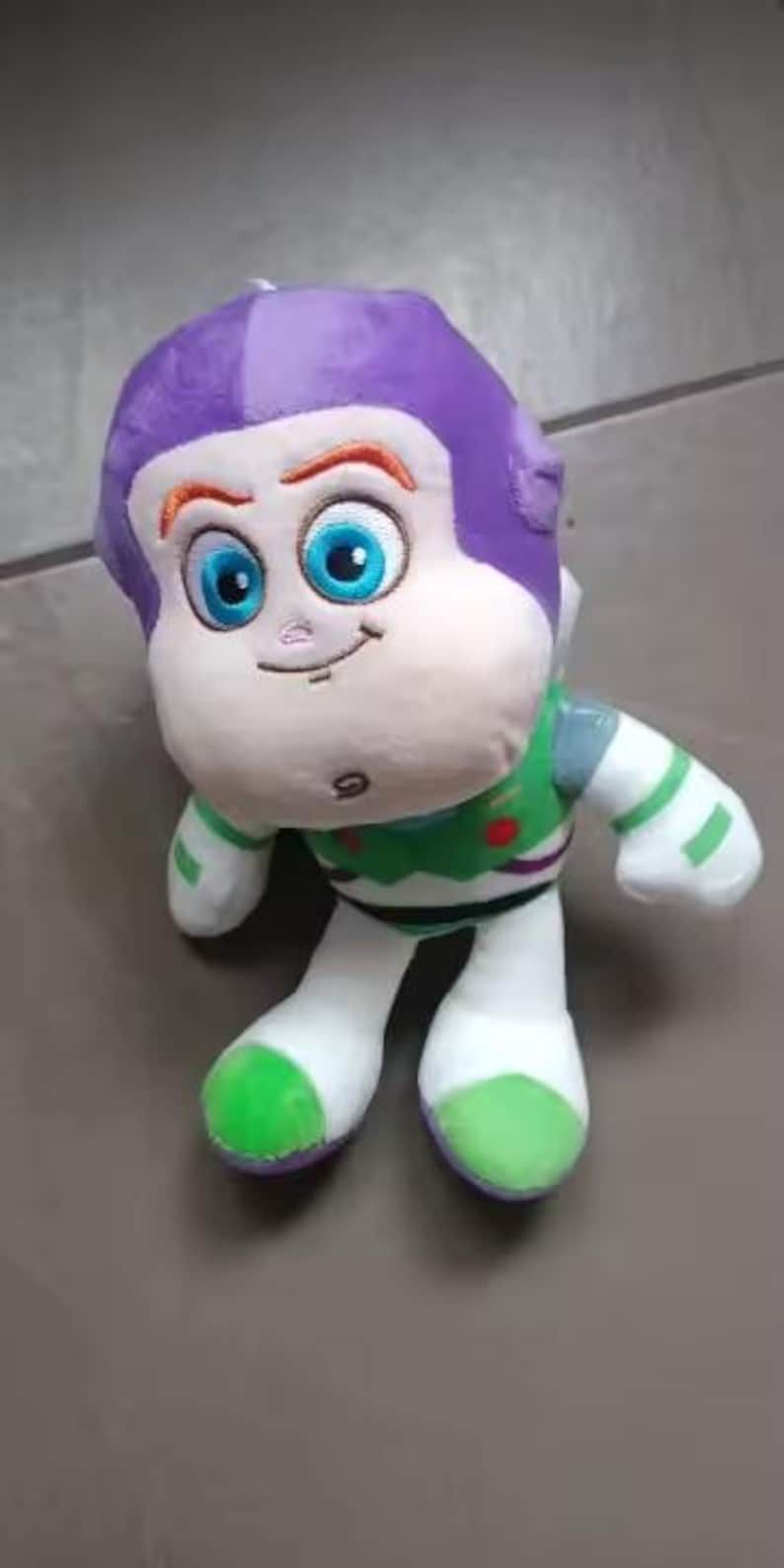 May include: A plush toy of the character Buzz Lightyear from the Toy Story franchise. The toy has a purple helmet, a white face, and a green and white spacesuit. The eyes are large and blue, and the toy is smiling.