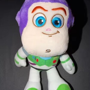 May include: A plush toy of Buzz Lightyear, featuring a purple head, large blue eyes, and a smiling expression. The toy has a white body with green and purple accents, and is designed to resemble the character from the animated film.