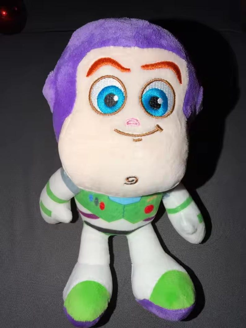 May include: A plush toy of Buzz Lightyear, featuring a purple head, large blue eyes, and a smiling expression. The toy has a white body with green and purple accents, and is designed to resemble the character from the animated film.