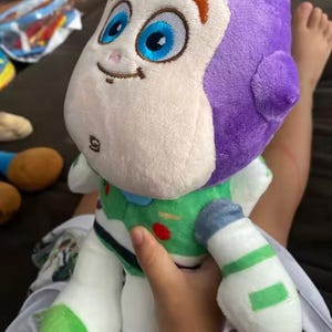 May include: A plush Buzz Lightyear doll, featuring a white face, purple head, and green and white spacesuit. The doll has large blue eyes, orange eyebrows, and a small smile. The toy is held by a person.