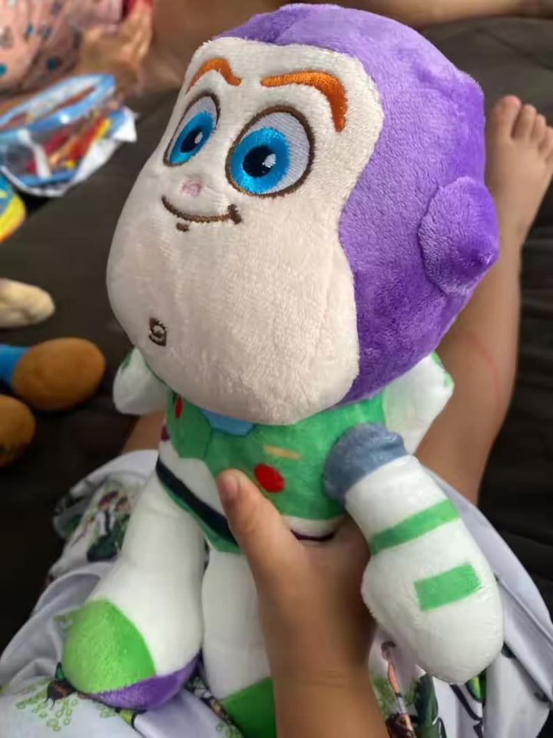 May include: A plush Buzz Lightyear doll, featuring a white face, purple head, and green and white spacesuit. The doll has large blue eyes, orange eyebrows, and a small smile. The toy is held by a person.