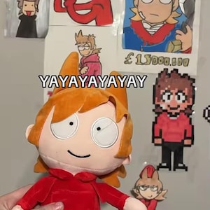 May include: A plush doll with orange hair and a red hoodie, black pants, and a smiling face. The doll is in front of drawings of the same character, including a "WANTED" poster. The text "YAYAYAYAYAY" is written on the wall.