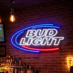 Peut inclure: Intérieur de bar avec enseigne au néon "Bud Light" bleue, blanche et rouge. Trois verres de bière sur le comptoir en bois, avec un bol de chips. En arrière-plan, un mur de briques, des bouteilles d'alcool et une télévision.
