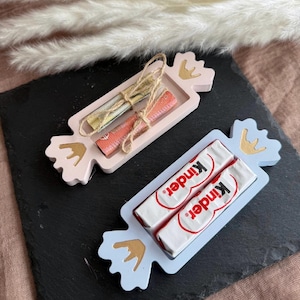 May include: Two candy-shaped trays, one pink and one blue, each holding items. The pink tray has rolled paper tied with twine. The blue tray holds two Kinder chocolate bars. Gold crown accents adorn the trays.