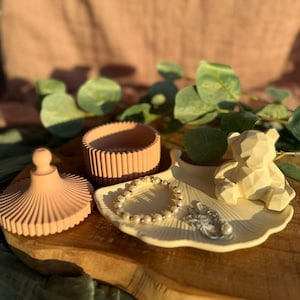 May include: A peach-coloured trinket box with a ribbed design, a small decorative bear, and a pearl bracelet rest on a wooden tray. The tray also holds a small, silver-coloured ring. Green leaves and a pink background complete the scene.
