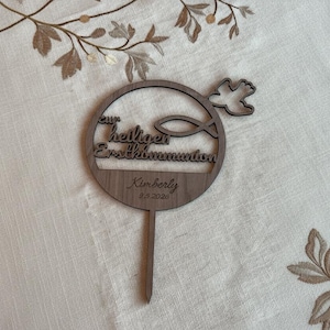 Puede incluir: Un adorno de pastel de madera marrón con el texto alemán "zur heiligen Erstkommunion" y un símbolo de pez. El adorno también incluye el nombre "Kimberly" y la fecha "9.5.2020". Una paloma está recortada en la parte superior. El adorno tiene un palo en la parte inferior.