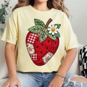 Puede incluir: Una camiseta amarillo claro con un gran diseño de fresa bordada. La fresa es roja con detalles de patchwork, hojas verdes y una margarita blanca. La camiseta es de estilo casual.