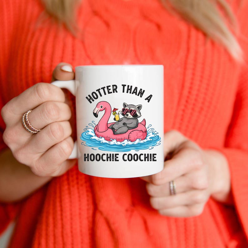 K&ouml;nnte beinhalten: Eine wei&szlig;e Keramik-Tasse mit schwarzem Henkel und dem Text "But First, Coffee" in fetter, schwarzer Schrift. Die Tasse steht vor einem verschwommenen Hintergrund, der eine gem&uuml;tliche Umgebung suggeriert.