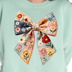 Op de afbeelding: Een lichtgroene sweatshirt met een grote, geborduurde strik. De strik is gemaakt van patchworkstof met bloempatronen in roze, oranje, blauw en geel. De strik is het centrale element van de sweatshirt.