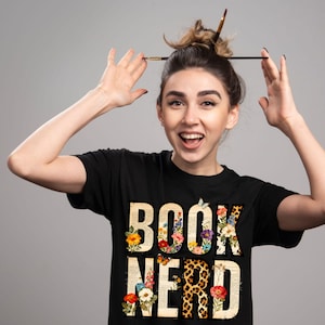 May include: Black t-shirt with the words "BOOK NERD" in a floral and leopard print design. The letters are filled with flowers, book pages, and leopard print. The person in the image is smiling and has a paintbrush in their hair.