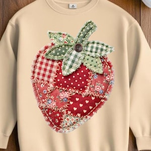 Puede incluir: Sudadera color crema con un gran diseño de fresa aplicado. La fresa está hecha de varios patrones de tela, incluyendo cuadros rojos, lunares, estampados florales y corazones blancos. Tiene hojas verdes y un botón marrón.