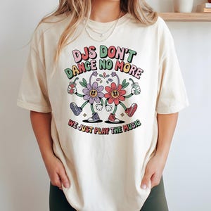 Può includere: T-shirt color crema con la scritta "DJs Don't Dance No More, We Just Play The Music". La maglietta presenta una grafica colorata di due fiori antropomorfi che tengono in mano delle bevande. I fiori indossano scarpe da ginnastica e hanno facce sorridenti.