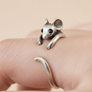 May include: A silver-colored ring featuring a 3D mouse design. The mouse has black eyes and is positioned as if it's climbing over the finger. The ring is adjustable and has a curved tail design.