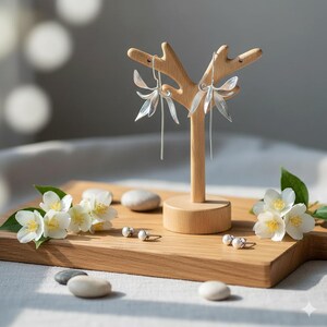 K&ouml;nnte beinhalten: Silberfarbene Blumenohrringe mit langen, baumelnden Elementen werden auf einem Holzschmuckst&auml;nder pr&auml;sentiert. Die Ohrringe sind mit kleinen Perlenohrsteckern und wei&szlig;en Blumen auf einem Holzbrett kombiniert.