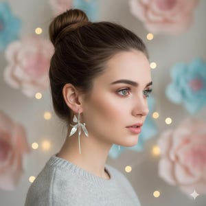 K&ouml;nnte beinhalten: Eine Frau tr&auml;gt silberne Ohrh&auml;nger mit floralem Design. Die Ohrringe sind lang und h&auml;ngen vom Ohr. Die Frau tr&auml;gt einen grauen Pullover und hat ihr Haar zu einem Dutt hochgesteckt. Der Hintergrund hat gro&szlig;e Papierblumen in Rosa und Blau.