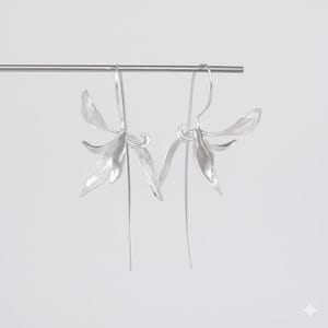 K&ouml;nnte beinhalten: Ein Paar silberfarbene Ohrringe mit floralem Design. Jeder Ohrring hat eine offene Bl&uuml;tenform mit mehreren Bl&uuml;tenbl&auml;ttern und einem langen, schlanken Stiel. Die Ohrringe sind an einem d&uuml;nnen, gebogenen Draht aufgeh&auml;ngt.