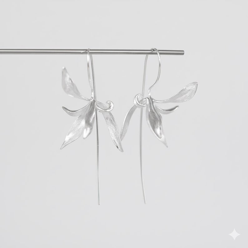 K&ouml;nnte beinhalten: Ein Paar silberfarbene Ohrringe mit floralem Design. Jeder Ohrring hat eine offene Bl&uuml;tenform mit mehreren Bl&uuml;tenbl&auml;ttern und einem langen, schlanken Stiel. Die Ohrringe sind an einem d&uuml;nnen, gebogenen Draht aufgeh&auml;ngt.