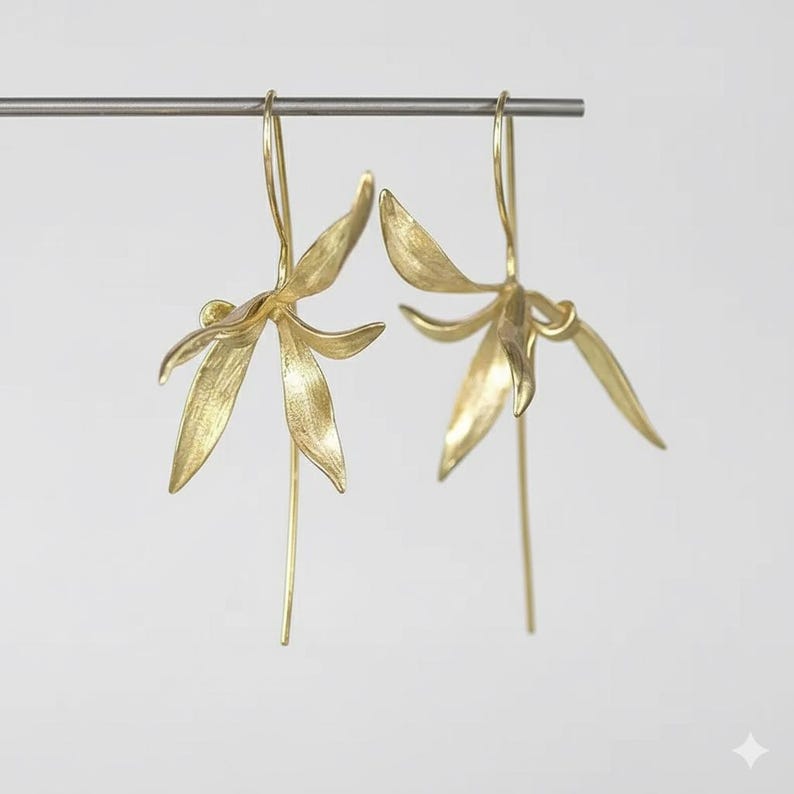 K&ouml;nnte beinhalten: Ein Paar goldfarbene Ohrringe in Bl&uuml;tenform. Jeder Ohrring hat ein zartes, bl&uuml;tenblattartiges Design mit einem langen, schlanken Tropfen. Die Ohrringe h&auml;ngen an einem d&uuml;nnen, silbernen Stab vor einem wei&szlig;en Hintergrund.