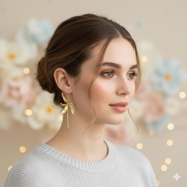 K&ouml;nnte beinhalten: Goldfarbene, baumelnde Ohrringe mit floralem Design. Die Ohrringe haben eine zarte, blattartige Form mit einem langen, schlanken Tropfen. Die Frau im Bild tr&auml;gt einen hellgrauen Pullover.