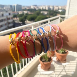 Pode incluir: Uma coleção de pulseiras coloridas em um pulso. Cada pulseira apresenta um cordão vibrante em tons de amarelo, vermelho, rosa, verde, azul e cinza, acentuado com uma conta decorativa dourada. As pulseiras são exibidas ao ar livre.