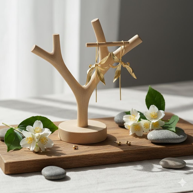 K&ouml;nnte beinhalten: Ein h&ouml;lzerner Schmuckst&auml;nder in Form eines Baumes, der goldene Blattohrringe pr&auml;sentiert. Die Ohrringe h&auml;ngen an einem Holzstab. Die Pr&auml;sentation befindet sich auf einem Holzbrett mit wei&szlig;en Blumen, gr&uuml;nen Bl&auml;ttern und kleinen grauen Steinen.
