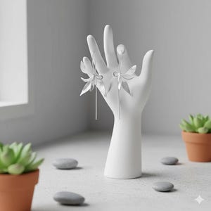 K&ouml;nnte beinhalten: Silberfarbene Blumenohrringe, pr&auml;sentiert auf einem wei&szlig;en, handf&ouml;rmigen Schmuckhalter. Die Ohrringe weisen filigrane, stilisierte Blumendesigns mit langen, schlanken Stiften auf. Der Hintergrund umfasst ein Fenster, kleine Topf-Sukkulenten und glatte graue Steine.