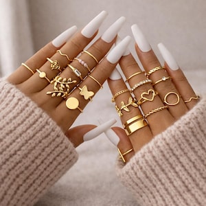 Może przedstawiać: Zestaw złotych pierścionków o różnych wzorach, w tym motyle, serca i motywy kwiatowe. Pierścionki są prezentowane na palcach z długimi, białymi paznokciami, na miękkim, neutralnym tle.