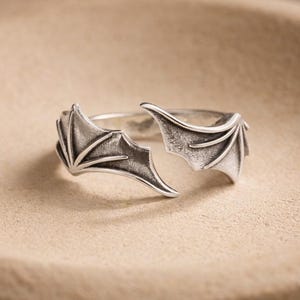 May include: A silver-toned ring with a unique design featuring stylized bat wings. The wings are detailed with textured veins and extend outwards, creating an open design. The ring is set against a neutral background.