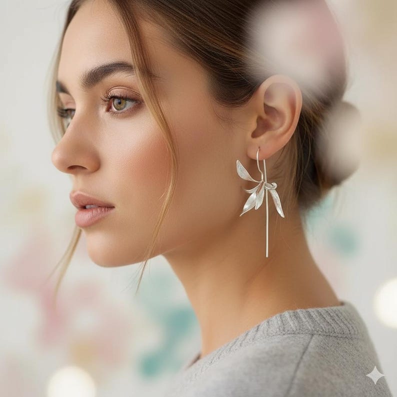 K&ouml;nnte beinhalten: Silberfarbene Blumenohrringe mit einem langen, schlanken Tropfen. Die Ohrringe haben ein zartes, blattartiges Design, das einen Hauch von von der Natur inspirierter Eleganz verleiht. Die Person tr&auml;gt einen grauen Pullover.