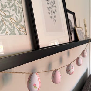 Może przedstawiać: Ozdobna girlanda wielkanocna z sześcioma materiałowymi jajkami. Jajka są w kolorze r&oacute;żowym i białym, z kwiatowymi i paskowanymi wzorami. Girlanda jest zawieszona na naturalnym sznurku i eksponowana na p&oacute;łce z oprawioną sztuką.