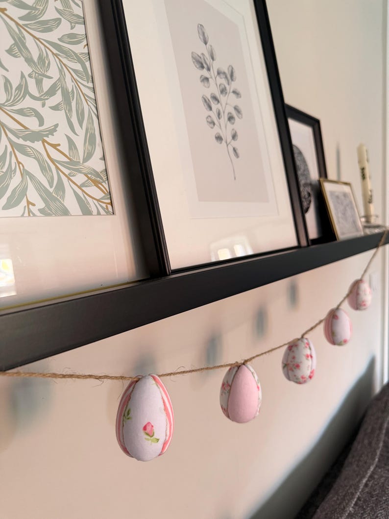 Może przedstawiać: Ozdobna girlanda wielkanocna z sześcioma materiałowymi jajkami. Jajka są w kolorze r&oacute;żowym i białym, z kwiatowymi i paskowanymi wzorami. Girlanda jest zawieszona na naturalnym sznurku i eksponowana na p&oacute;łce z oprawioną sztuką.