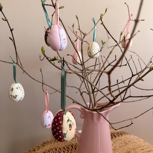 May include: A pink jug displays branches decorated with fabric Easter egg ornaments. The eggs have floral and polka dot patterns in pink, white, and burgundy, suspended by ribbons. The arrangement is on a woven surface.