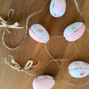 Może przedstawiać: Ozdobna girlanda wielkanocna z jajkami. Jajka wykonane są z tkaniny z kwiatowymi i paskowymi wzorami w kolorze r&oacute;żowym, białym i jasnoniebieskim. Girlanda jest nawleczona na sznurek i zawiera małe, kremowe kokardki.