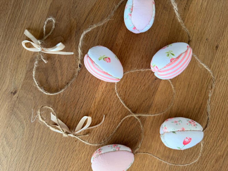 Może przedstawiać: Ozdobna girlanda wielkanocna z jajkami. Jajka wykonane są z tkaniny z kwiatowymi i paskowymi wzorami w kolorze r&oacute;żowym, białym i jasnoniebieskim. Girlanda jest nawleczona na sznurek i zawiera małe, kremowe kokardki.