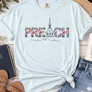 以下が含まれることがあります： 花柄で描かれた「PREACH」の文字がプリントされたライトブルーの半袖Tシャツ。文字の「A」の部分は教会のイラストになっています。クルーネックで、右下の裾が結ばれたデザインの衣類です。