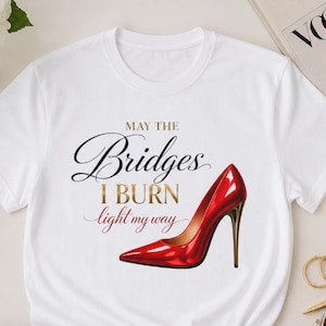 May include: White t-shirt featuring a red high-heeled shoe graphic and the text "May the Bridges I Burn light my way." The shirt is styled with black shorts, a black purse, and a Vogue magazine.