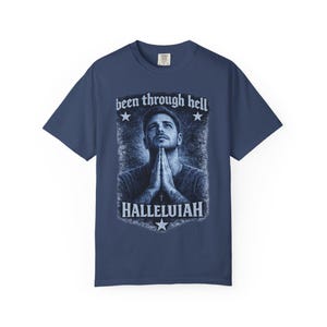 May include: Navy blue t-shirt featuring a graphic of a man with hands clasped in prayer. The text "been through hell" and "Hallelujah" are above and below the image, with stars and a cross accenting the design. The shirt is a classic crew neck.