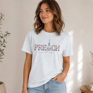 以下が含まれることがあります： 胸元に「PREACH」の文字がプリントされたライトブルーの半袖Tシャツ。文字はダークな花柄で埋め尽くされており、「A」の文字は教会の尖塔の線画に置き換えられ、その下に装飾的なラインが引かれています。